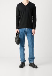 Pullover nero con scollo a V, jeans in denim blu, stivali in pelle nera e borsa nera, con dettagli del logo visibili sul pullover.