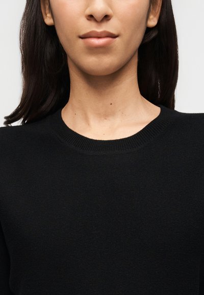 Γυναίκα που φοράει απλό μαύρο πουλόβερ με στρογγυλή λαιμόκοψη, εμφανίζοντας το λαιμό και το πάνω μέρος του στήθους με φόντο έναν απλό τοίχο.