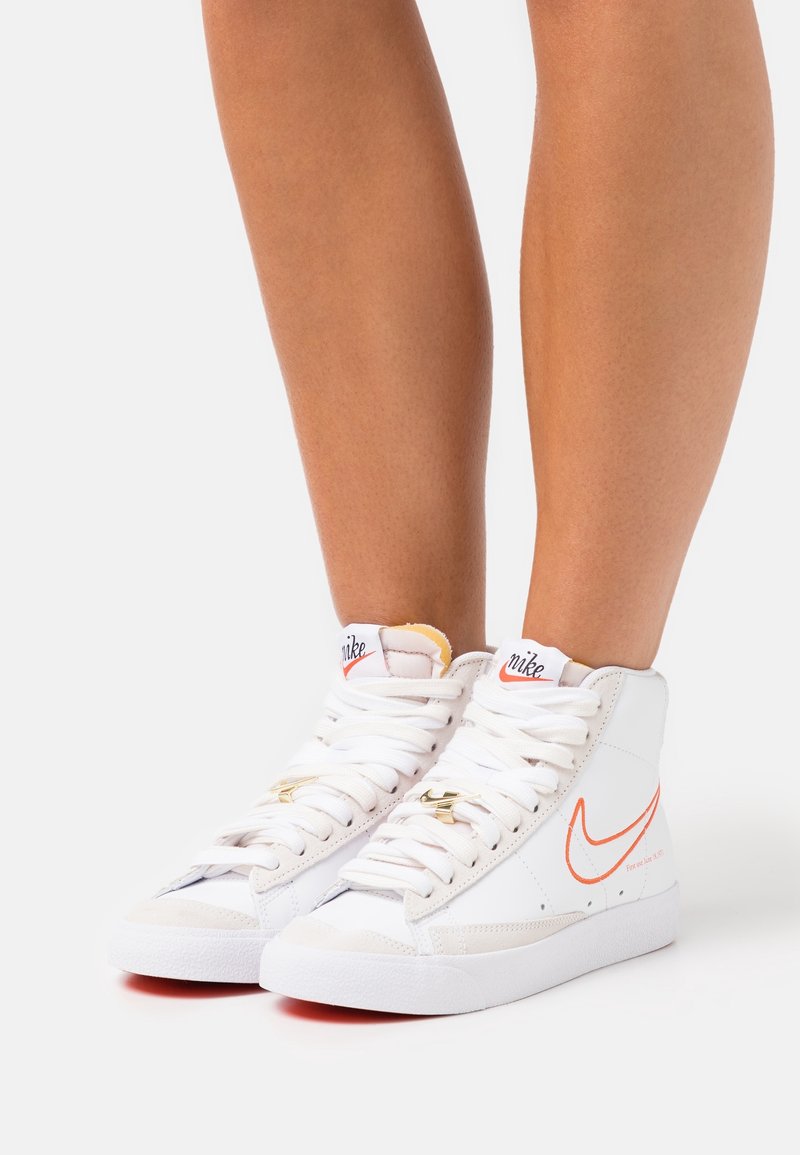 Gambe nude che indossano sneakers Nike bianche alte con il logo arancione dello swoosh e lacci bianchi, in piedi su uno sfondo bianco.