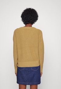 Senfgelber Strickpullover mit lockerer Passform und geripptem Material, kombiniert mit einem dunklen Jeansrock mit aufgesetzten Taschen.