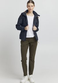 Giacca blu navy con pois bianchi, zip frontale, cappuccio e dettagli marroni, abbinata a pantaloni verde oliva e sneaker bianche.