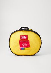 Yellow mesh travel bag with a black trim, featuring a red label displaying product details. Round shape with a top handle for easy carrying.