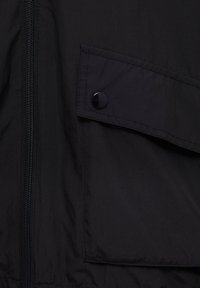 Veste noire légère avec une fermeture éclair à l'avant, poche à rabat avec bouton-pression, et texture lisse. Design minimaliste sans motifs visibles.
