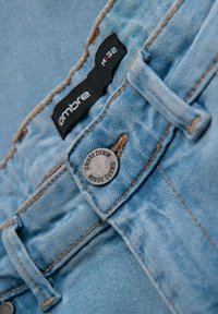 Jeans de mezclilla azul claro con una etiqueta negra que dice "ombre M32", con un botón de metal grabado con "OMBRE DENIM". Costuras en contraste visibles.