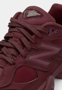 Maroon New Balance 9060 sneaker met textuur materialen, dikke veters, en een zichtbaar logo met een oogje-ontwerp. Gebogen vorm met omliggende accenten.