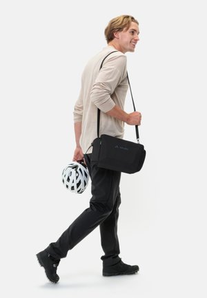 Borsa a tracolla nera con una tracolla regolabile, materiale testurizzato e un piccolo dettaglio verde. Un casco da bicicletta bianco è tenuto nell'altra mano.
