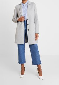 Manteau gris clair en mélange de laine, au design ajusté, à simple boutonnage avec deux boutons et poches latérales, associé à une chemise rayée et des talons hauts.
