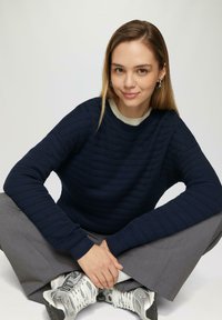 Marineblauwe geribbelde gebreide trui met een ronde halslijn, gecombineerd met grijze wijde broekspijpen en metallic sneakers, met textuurdetails in de stof.