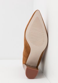 Chaussure en daim marron à bout pointu, posée en biais contre un mur blanc, montrant sa semelle beige texturée et son talon bloc avec une semelle en caoutchouc antidérapante.