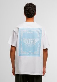 Hombre de espaldas vistiendo una camiseta blanca con un gran gráfico cuadrado azul y texto y patrones decorativos blancos en la parte posterior.