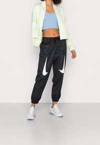 Kvinna som bär svarta Nike-joggers med vita swoosh-loggor, en ljusblå kortärmad tanktop, en ljusgrön zip-up jacka och vita sneakers.
