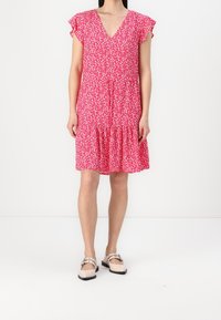 Rosa geblümtes Kleid mit kurzen, gerafften Ärmeln, V-Ausschnitt und Stufenrock. Der Stoff hat eine leichte, strukturierte Optik. Kombiniert mit flachen Schuhen.