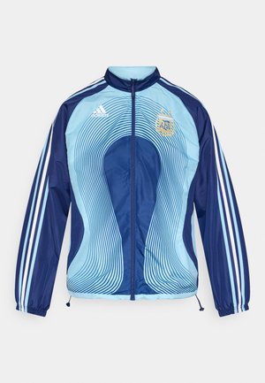 Lyseblå og marineblå vindjakke med glidelås foran, med buede linjemønstre, tre hvite striper på ermene, og en brodert logo.