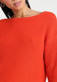 Jasnopomarańczowy sweter z dzianiny z szerokim dekoltem w kształcie łodzi, luźnym krojem i teksturowanym materiałem. Posiada długie rękawy i prążkowane detale.