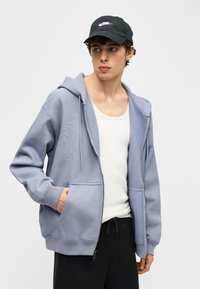 Νέος άνδρας που φοράει ανοιχτό μπλε φούτερ με φερμουάρ, λευκό φανελάκι, μαύρο παντελόνι και μαύρο καπέλο Nike, στέκεται με το ένα χέρι στην τσέπη.