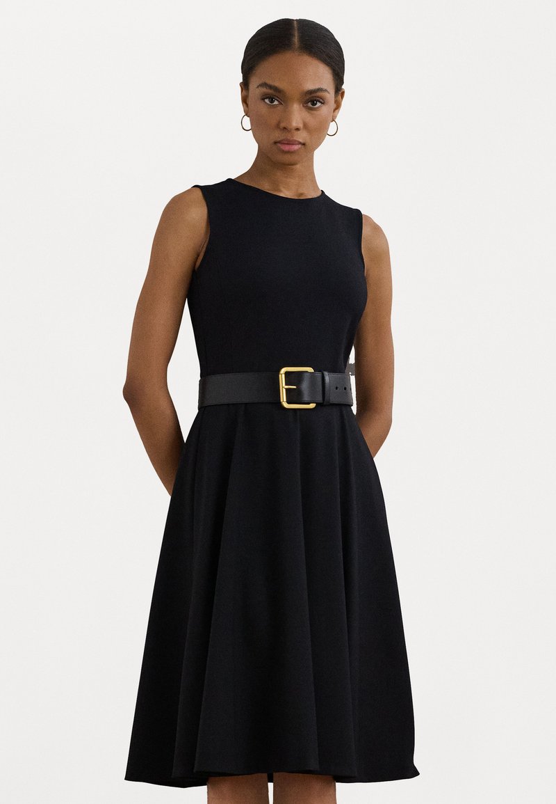 Melnas bez piedurknēm kleita, izgatavota no gludas auduma, ar izvērstu svārku un pieguļošu jostasvietu, kas akcentēta ar platu melnu jostu un zelta sprādzi.