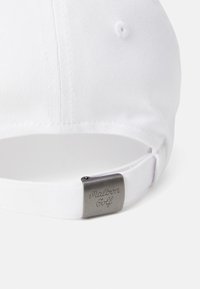 White cap with structured design, adjustable strap with metal clasp featuring "Malbon Golf" engraving, smooth texture, and stitched detailing.