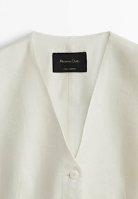 Gros plan d'un vêtement beige à col en V avec un seul bouton et une étiquette noire indiquant "Massimo Dutti, Made in Morocco."