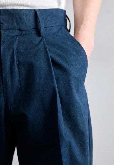 Pantalons bleu marine en coton avec des plis à l'avant, une texture lisse et des poches latérales ; ceinture ajustée et détails de couture minimalistes.