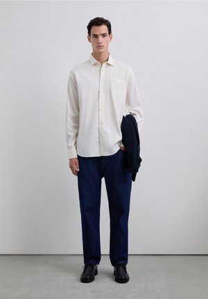 Lichtcrème shirt met lange mouwen, knoopsluiting en een borstzak, gecombineerd met donkerblauwe jeans en zwarte schoenen, terwijl hij een marineblauwe blazer vasthoudt.