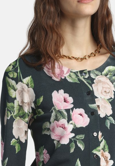 Cardigan vert foncé avec un motif floral de fleurs roses et crème, col côtelé et boutons noirs. Orné d'un collier chaîne doré.
