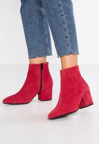 Bottes cheville en daim rouge avec un bout pointu, une fermeture éclair latérale et un talon épais. Portées avec un jean denim bleu raccourci présentant des bords effilochés.