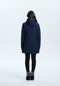 Giacca impermeabile blu navy con cappuccio, vita stretta e design con rétro più lungo. Indossata con leggins neri e stivali neri con bordo di pelliccia.