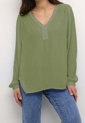 Kvinde iført en lysegrøn bluse med lange ærmer og V-hals samt blå jeans, stående med hænderne afslappet langs siden mod en ensfarvet baggrund.