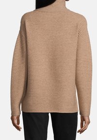 Beige ribbat stickad tröja med hög krage och lös passform, har en texturerad yta och sänkta axlar, tillsammans med mörka byxor.