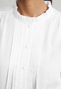 Weißes, strukturiertes Blusenshirt mit Rüschensaum, vertikalen Falten und Knopfverschlüssen vorne. Glattes Material mit leichtem Fall.