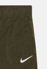 Olivgröna shorts i texturerat tyg, med en sidoficka och en vit Nike-logotyp omgiven av guld på sidan.