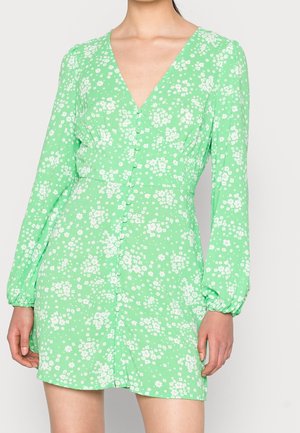 Kobieta w jasnej zielonej sukience w kwiaty z długimi rękawami, zapinanej na guziki z przodu, z dekoltem w serek, sfotografowana od szyi do połowy ud.