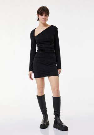 Młoda osoba o krótkich włosach, ubrana w dopasowaną czarną sukienkę na jedno ramię i czarne kozaki do kolan na białym tle.