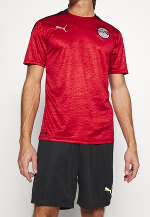 Mies, joka pukeutunut punaiseen jalkapallopaitaan, jossa on mustia geometrisia kuvioita ja Egyptin jalkapalloliiton logo, yhdistettynä mustiin Puma-housuihin.