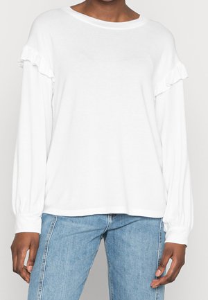 Nainen pukeutuneena valkoiseen pitkähihainen paitapuseroon, jossa röyhelöyksityiskohta rinnalla, ja vaaleansiniset korkeavyötäröiset farkut, seisomassa yksiväristä taustaa vasten.