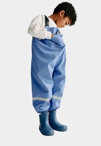 Blå vandtætte overalls med elastiske manchetter, refleksstriber og lommer, båret over blå gummistøvler. Mønstrefri, glat tekstur.