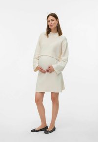 Lichtbeige zwangerschapsjurk met een ribbeltextuur, lange mouwen en een cropped top. Gdragen met zwarte gedecoreerde ballerina's.