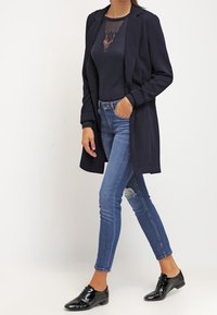 Long blazer marine sur un haut ajusté marine, associé à un jean bleu déchiré et des chaussures noires en cuir verni. Tissu lisse, coupe cintrée.