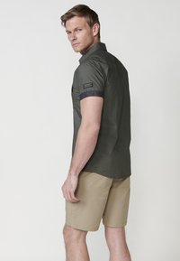 Camisa de manga corta en color verde oliva, hecha de tela ligera; presenta un puño en azul oscuro y un bolsillo en el pecho con detalles en texto, combinada con pantalones cortos beiges.