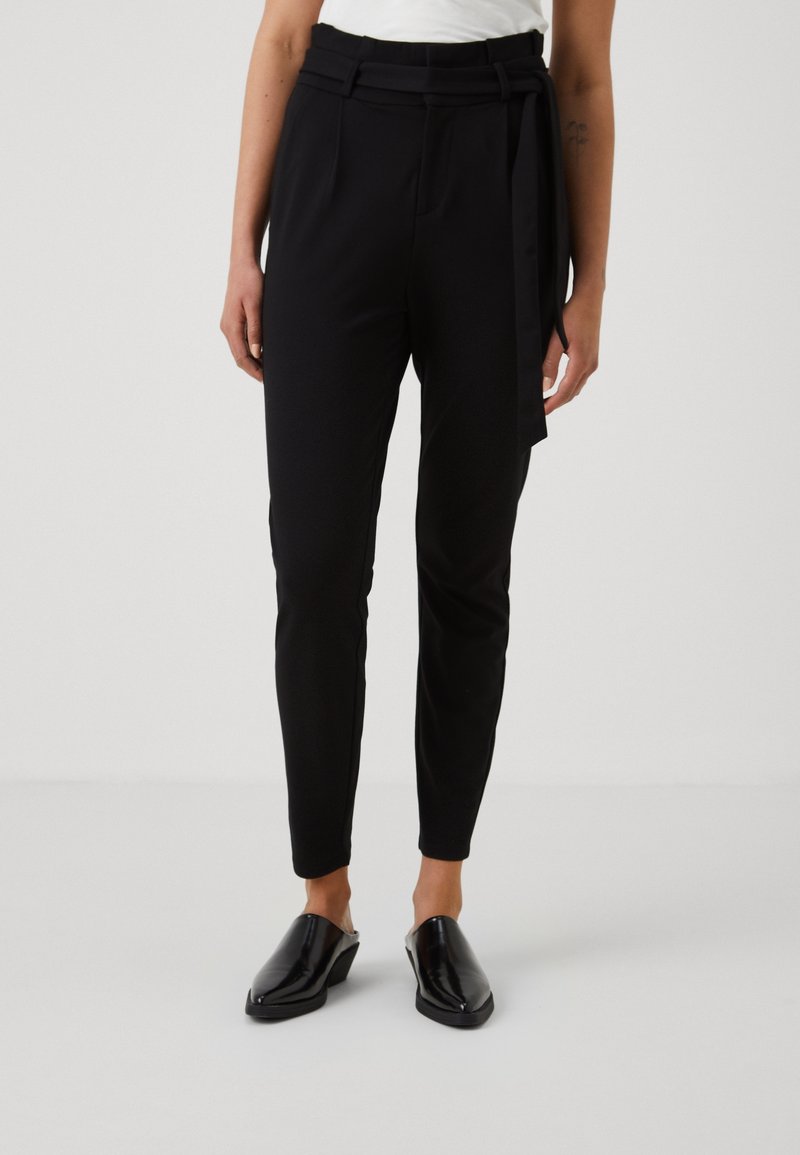 Femme portant un pantalon noir taille haute fuselé avec une ceinture en tissu nouée à la taille, des chaussures noires à talons sans lacets et un haut blanc.