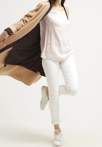 Lange bruine en tan jas met contrasterende manchetten, lichtroze top, witte jeans en witte sneakers met gele details. Model in dynamische pose.