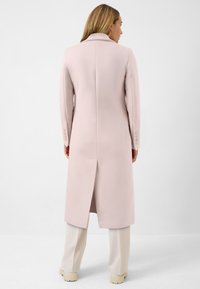 Ljusrosa lång rock med en strukturerad design, slagkrage och en mitt bak-ventil. Har knappar som dekoration på ärmarna. Neutralt färgade byxor.