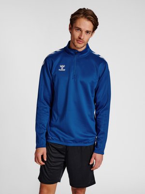 Joven con un pulóver deportivo azul con cremallera hasta la mitad y rayas blancas en forma de cheurón en los hombros y pantalones cortos deportivos negros, de pie frente a un fondo liso.