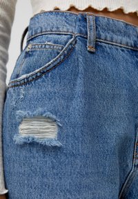 Siniset farkut korkealla vyötäröllä, joissa on kulutettuja yksityiskohtia ja revitty reikä reidessä, joiden kanssa on oranssia ommelta korostamassa ilmettä.