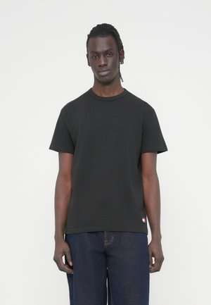 Jeune homme à la peau foncée avec de courts dreadlocks, portant un t-shirt noir uni à manches courtes et un jean bleu foncé, debout devant un fond blanc.