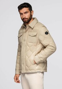 Uomo con capelli corti e scuri e barba, che indossa un giubbotto trapuntato beige con tasche a patta, in piedi di fronte a uno sfondo chiaro e uniforme.