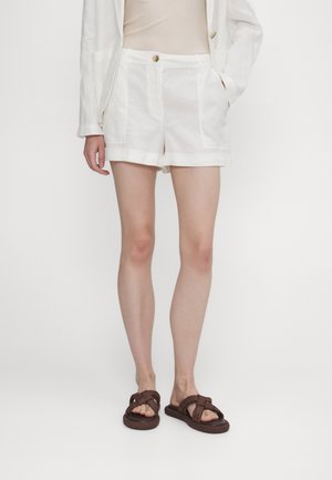 Personne portant un short blanc ajusté, un haut beige, un blazer blanc et des sandales tressées marron, debout devant un fond blanc uni.