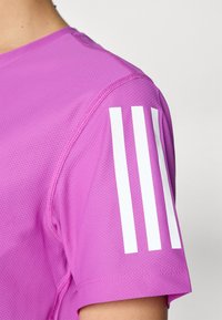 Krátky rukáv atletického trička vo výraznej ružovej farbe, vyrobené z textúrovanej látky, s troma bielymi pruhmi na ľavom rukáve.