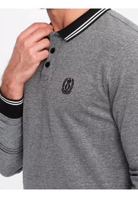 Polo gris avec un col classique, trois boutons noirs et un petit logo brodé. Manches avec poignets rayés noir et blanc.