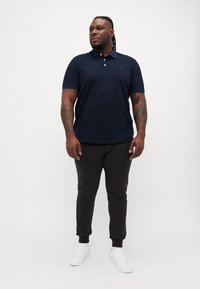 Marineblå polo-skjorte med krage, to knapper og korte ermer, kombinert med svarte joggebukser og hvite joggesko. Glatt stoff, enkelt design.